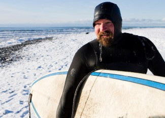 An Alaskan surfer walks over a snowy beach after a winter surf s